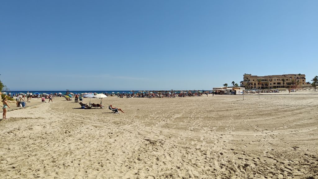 Wide stretch of sand at La Zenia beach on a sunny day, with sunbathers in the distance and seafront buildings on the headland