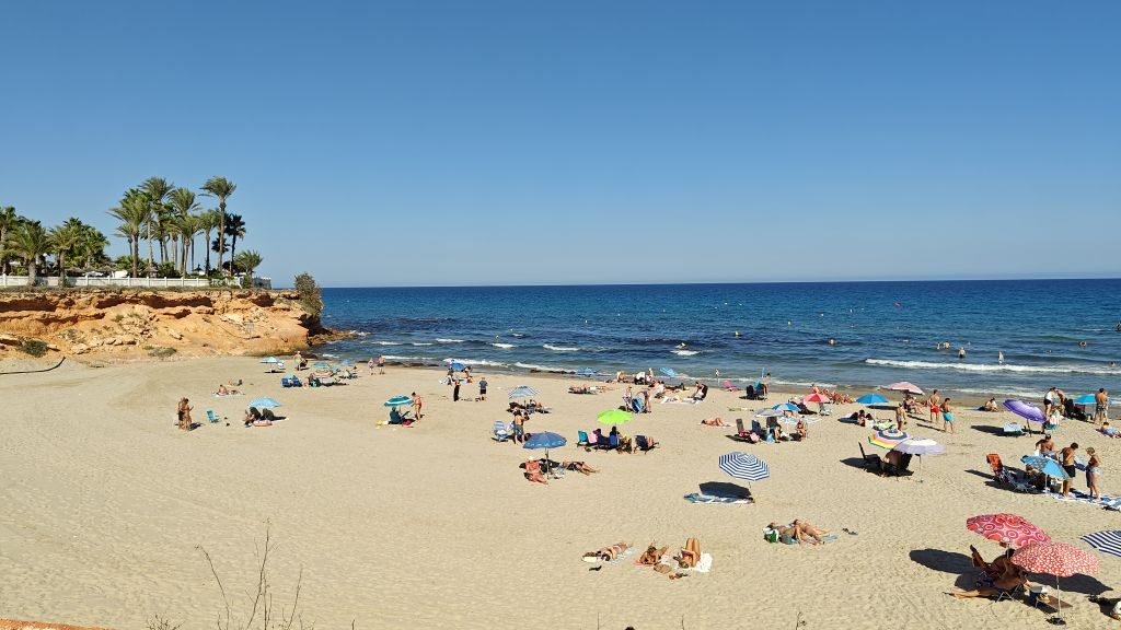 Colourful parasols and sunbathers on La Zenia beach, with a palm-topped sandstone headland behind
