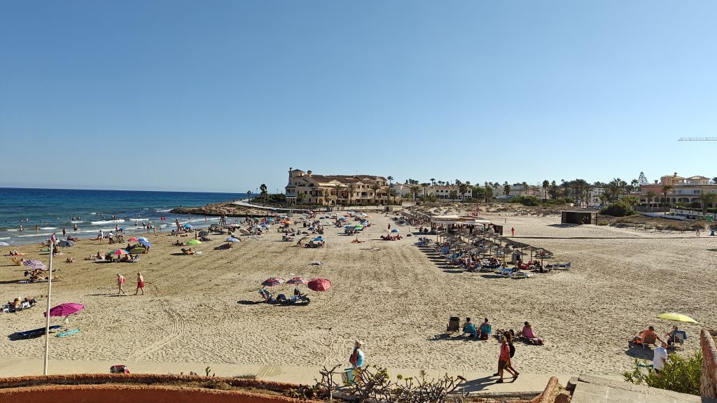 A busy La Zenia beach on a sunny day, looking along the bay towards the seafront buildings