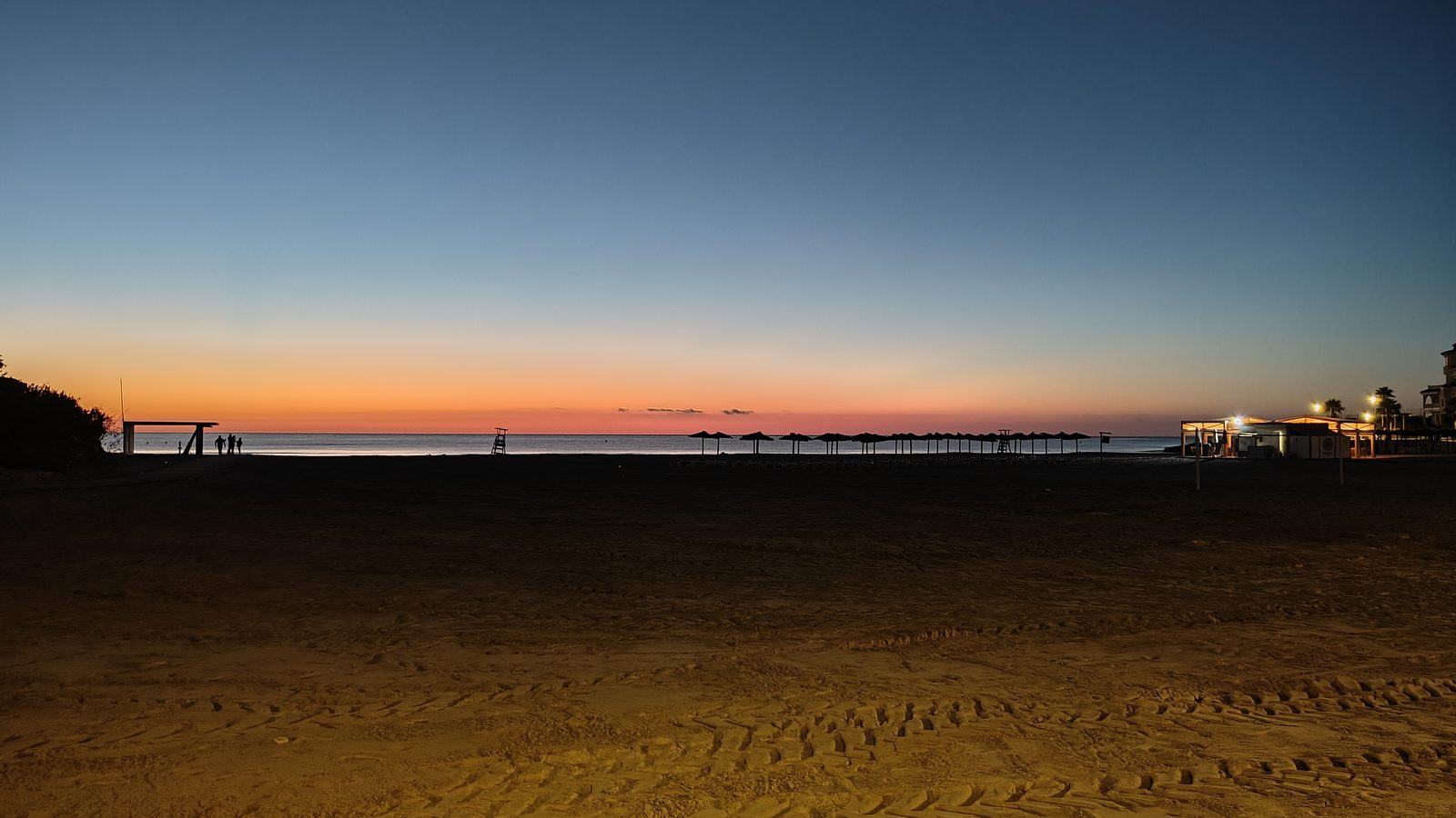 Rows of beach parasols and a lit beach bar along La Zenia beach before sunrise