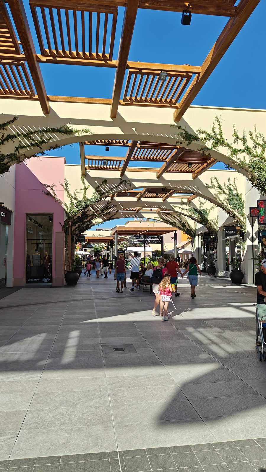 Open-air walkway at Zenia Boulevard shaded by a timber pergola and climbing plants